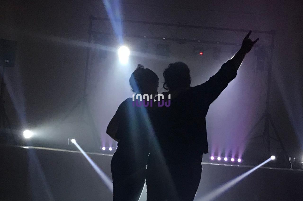 Deux personnes de dos dans une salle sombre, éclairées par des projecteurs et des lumières de scène.