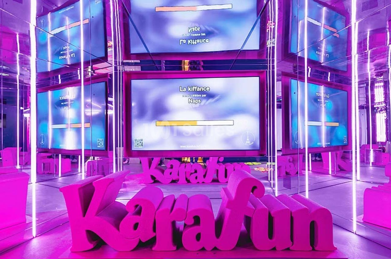 Salle de karaoké avec écrans affichant des paroles de chanson, éclairée par des lumières roses et violettes.