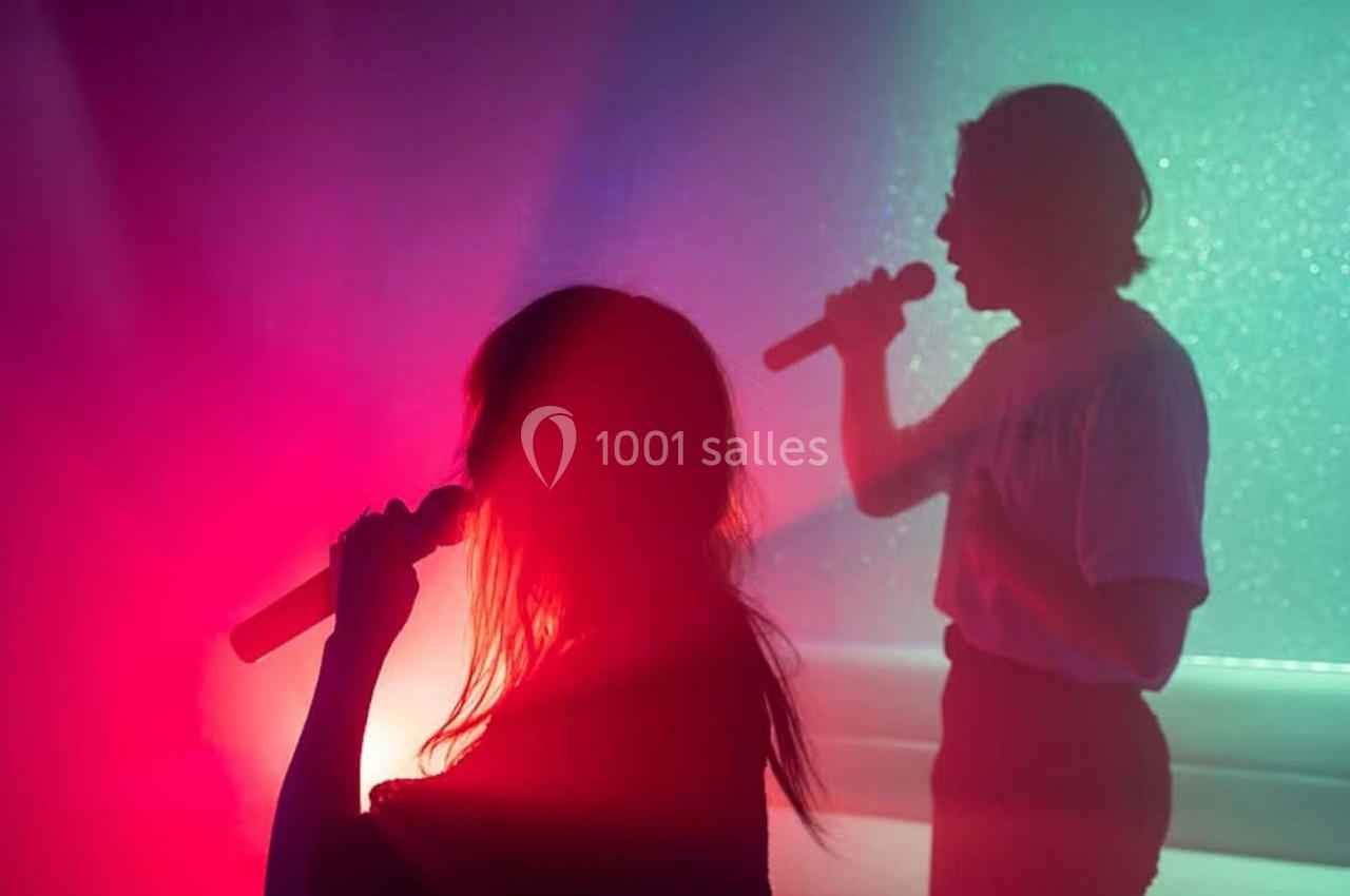 Deux personnes chantent au micro dans une ambiance lumineuse colorée avec des tons rouges et verts.