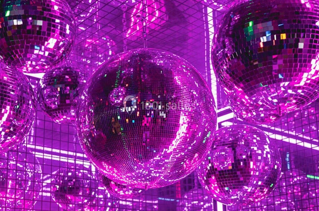 Boules à facettes roses suspendues dans une pièce aux murs recouverts de miroirs et éclairée par des néons violets.