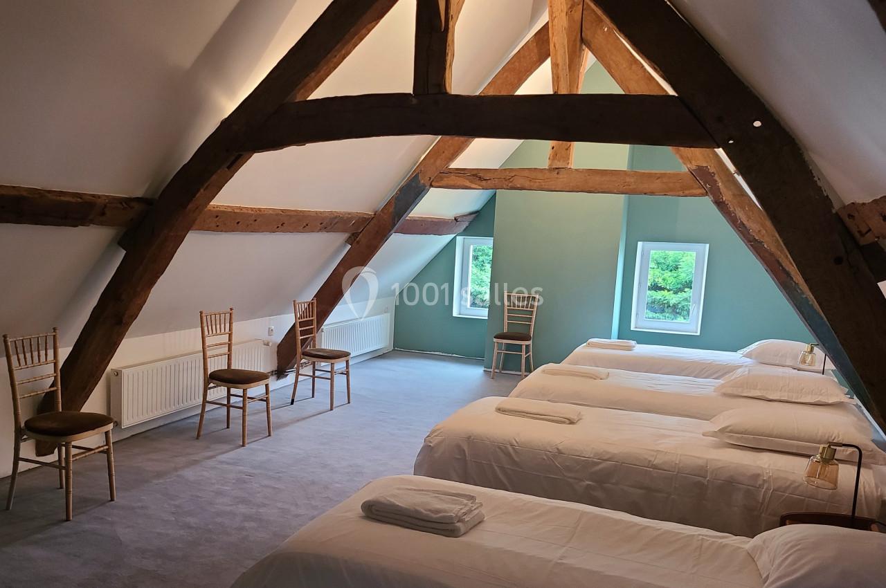 Salle mansardée avec poutres apparentes, quatre lits simples alignés, chaises en bois et fenêtres donnant sur un paysage…