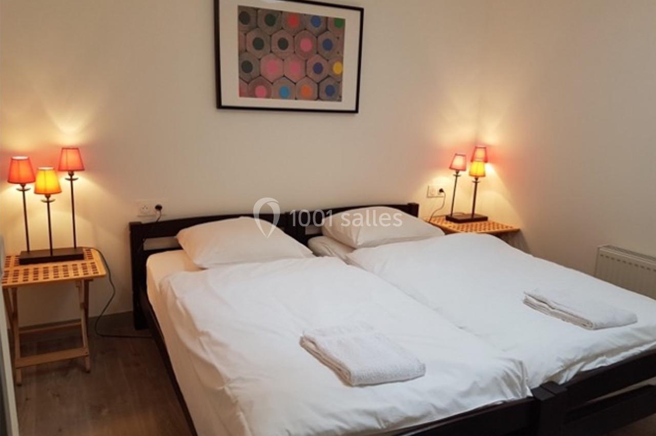 Deux lits simples avec draps blancs, tables de chevet en bois, lampes allumées et tableau coloré au mur.