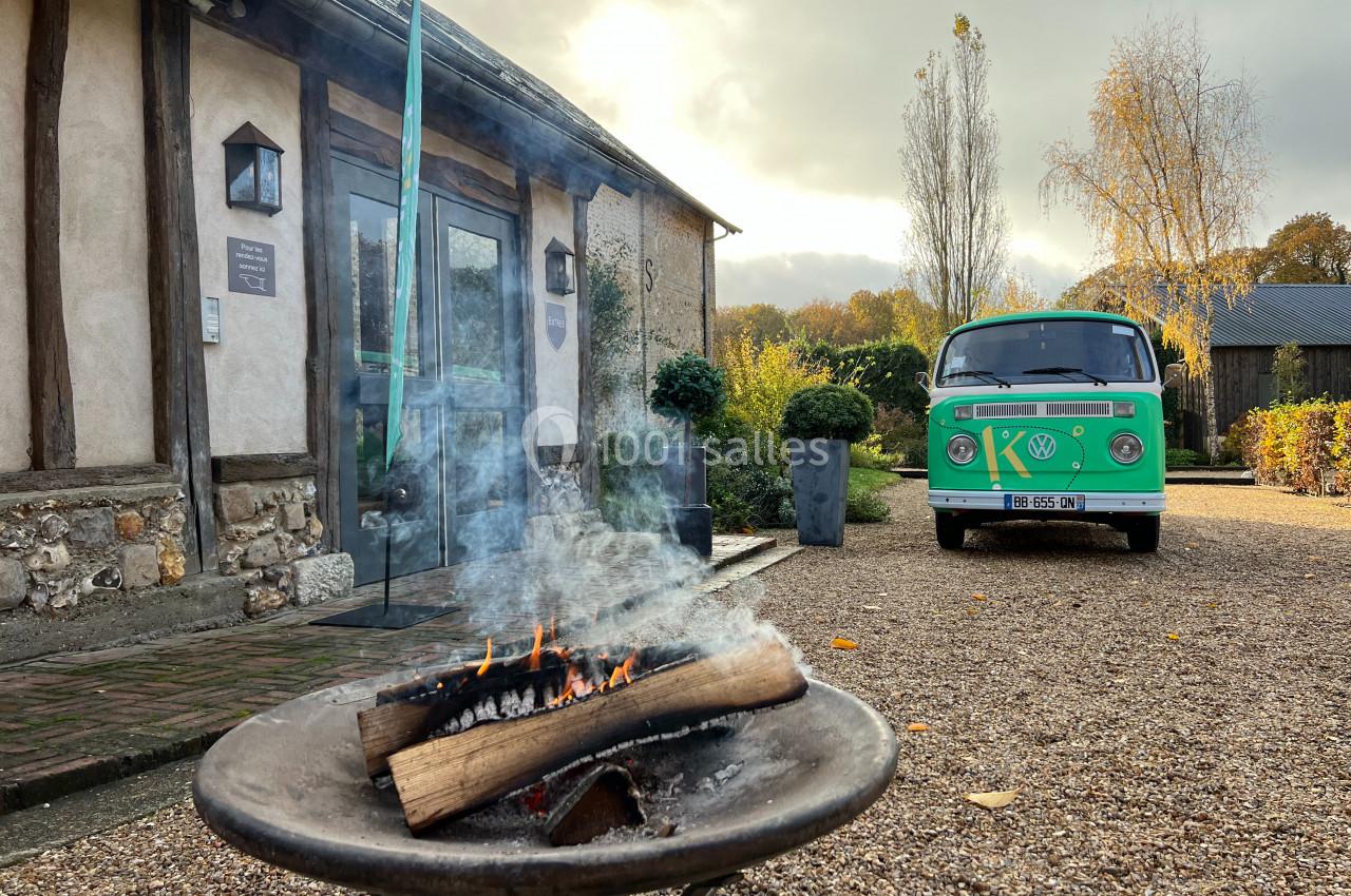 Brasero allumé devant une maison en pierre et bois, avec un van vert garé sur une allée gravillonnée.