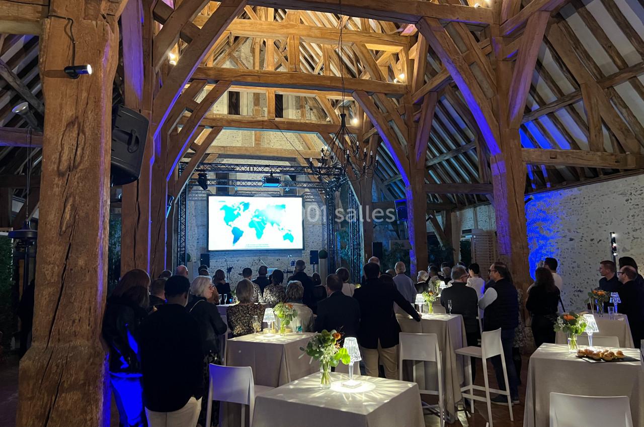 Salle en bois avec des poutres apparentes, des invités debout regardant une présentation projetée sur un écran.