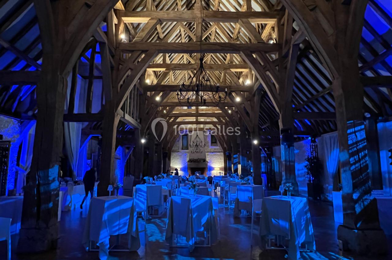Salle de réception avec charpente en bois apparente, éclairage bleu et tables dressées pour un événement.