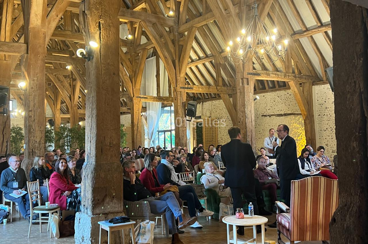 Un groupe de personnes assiste à une conférence dans une grande salle avec poutres apparentes et éclairage chaleureux.