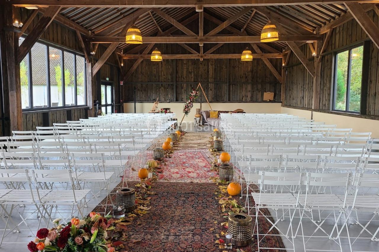 Salle de réception en bois décorée pour une cérémonie, avec chaises blanches alignées, tapis, citrouilles et fleurs.
