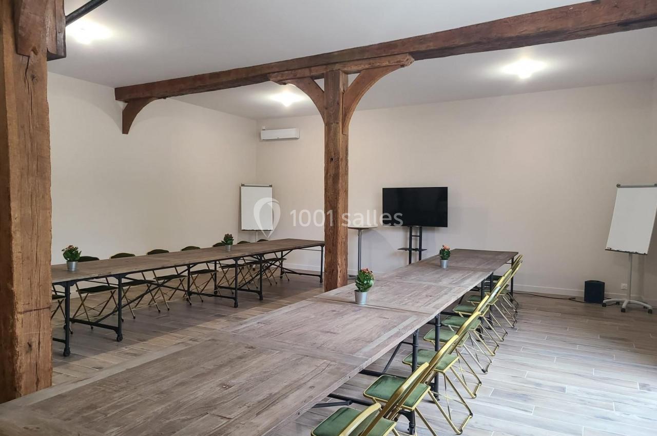 Salle de réunion lumineuse avec tables en bois disposées en U, chaises pliantes, écran, paperboards et poutres apparentes.