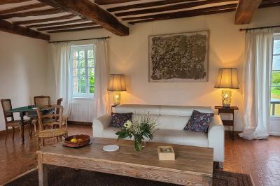 Salon lumineux avec poutres apparentes, canapé blanc, table basse en bois, lampes allumées et vue sur jardin.