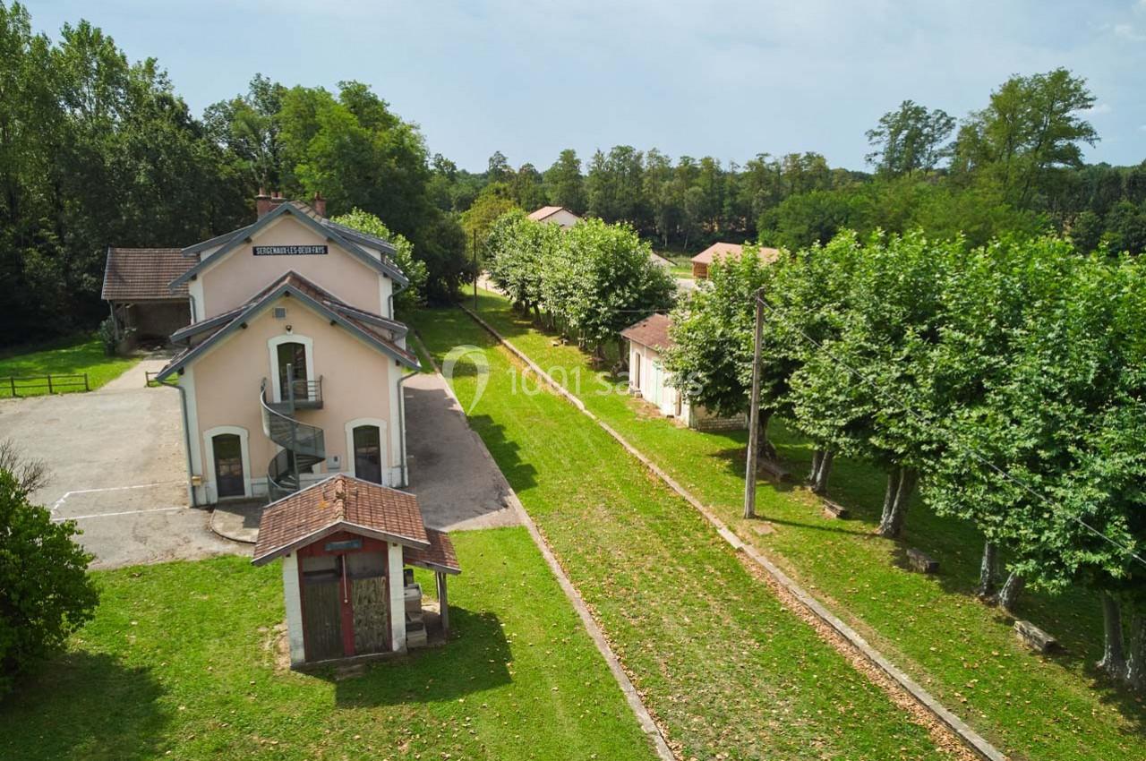 Ancienne gare entourée d'arbres, avec voie ferrée désaffectée et bâtiments annexes dans un cadre verdoyant.