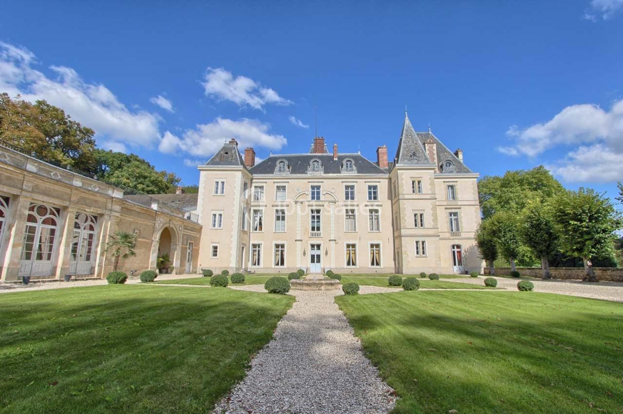 Façade d'un château entouré de pelouses bien entretenues, avec un ciel bleu et quelques nuages en arrière-plan.