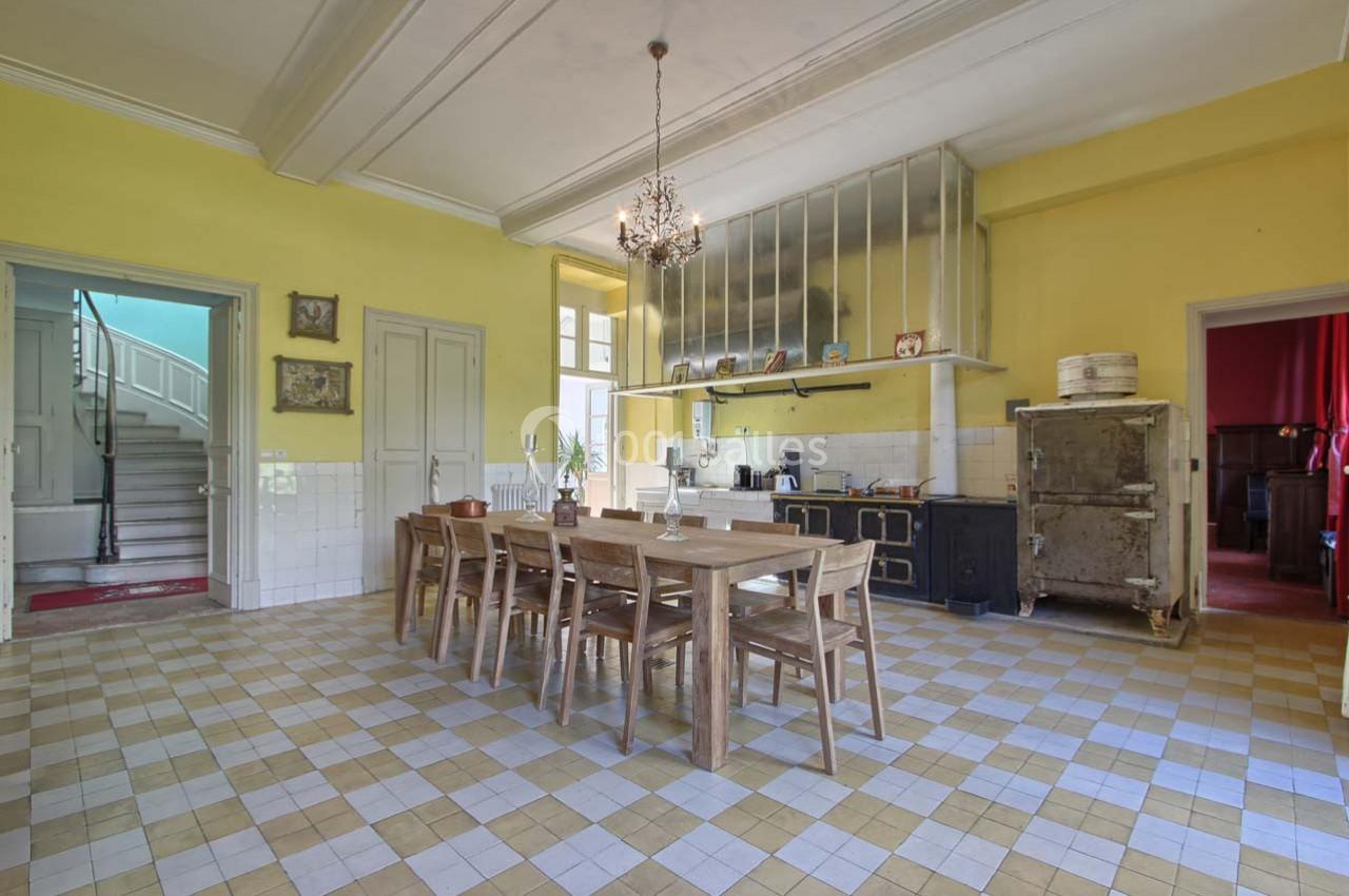 Grande cuisine lumineuse avec murs jaunes, table en bois massif, carrelage à motifs et escalier visible en arrière-plan.