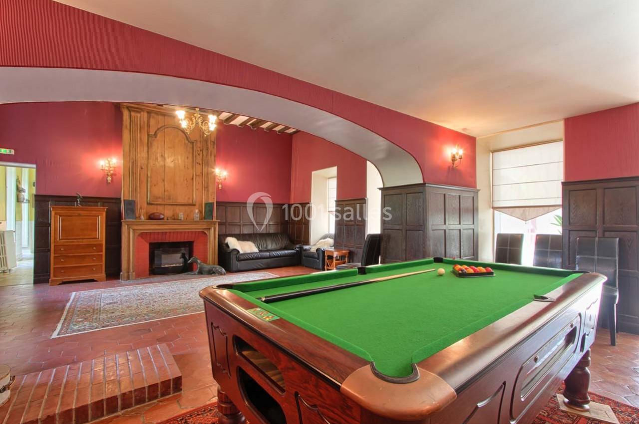 Salle avec une table de billard au premier plan, un canapé près d'une cheminée et des murs ornés de boiseries.