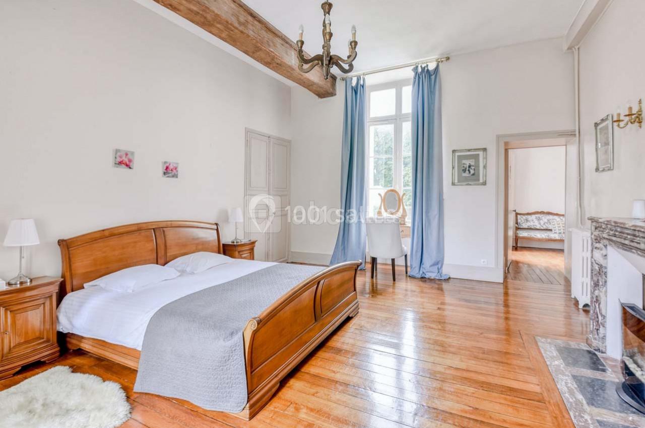 Chambre lumineuse avec lit double en bois, parquet, cheminée, poutre apparente et rideaux bleus devant une grande fenêtre.