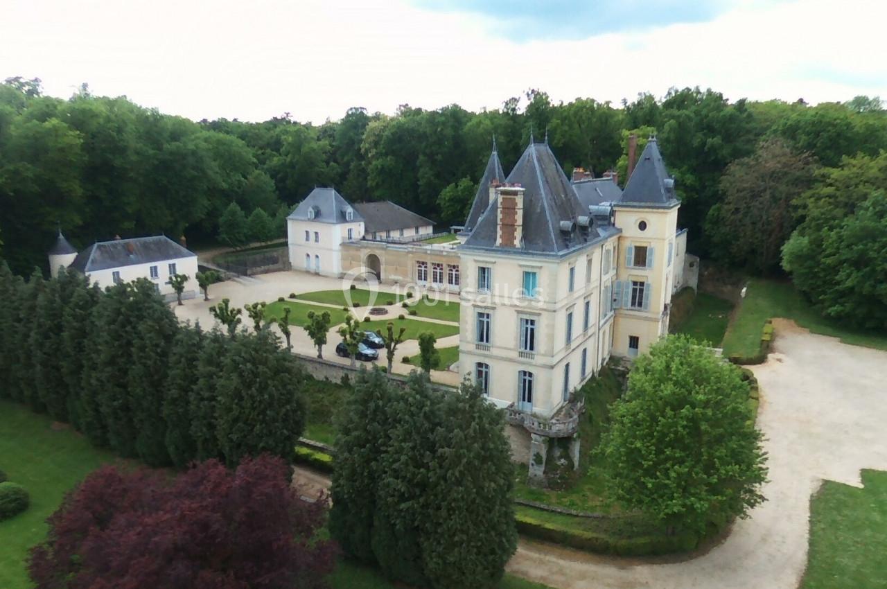 Vue aérienne d'un château entouré de jardins, d'allées gravillonnées et d'une forêt dense en arrière-plan.