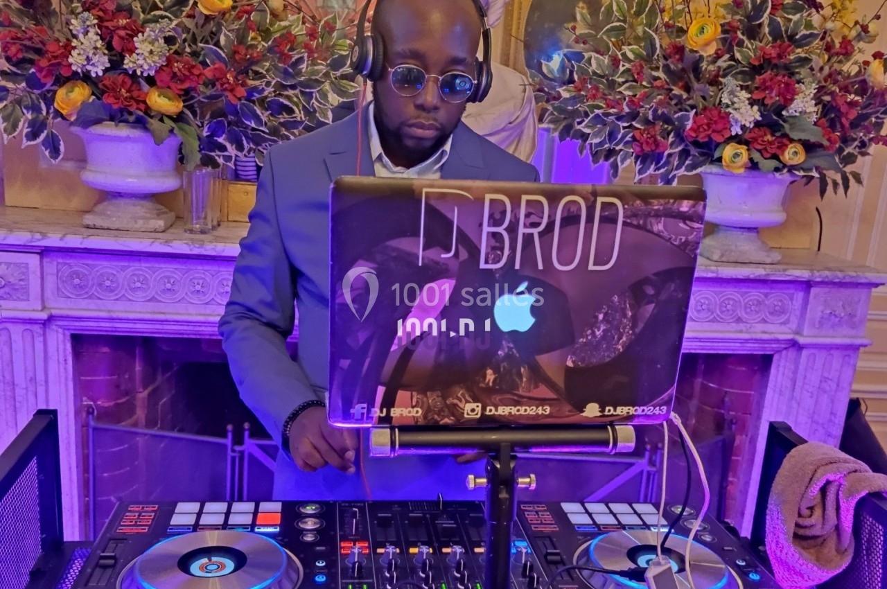 Un DJ mixant de la musique sur une console, devant un décor floral et une cheminée éclairée en violet.