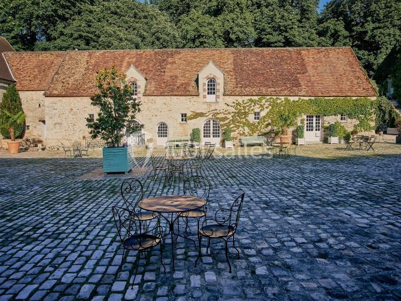 Cour pavée avec des tables et chaises en fer forgé devant un bâtiment en pierre aux toits de tuiles rouges.