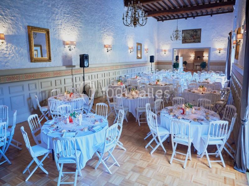 Salle de réception élégante avec tables rondes dressées, chaises blanches et éclairage chaleureux.