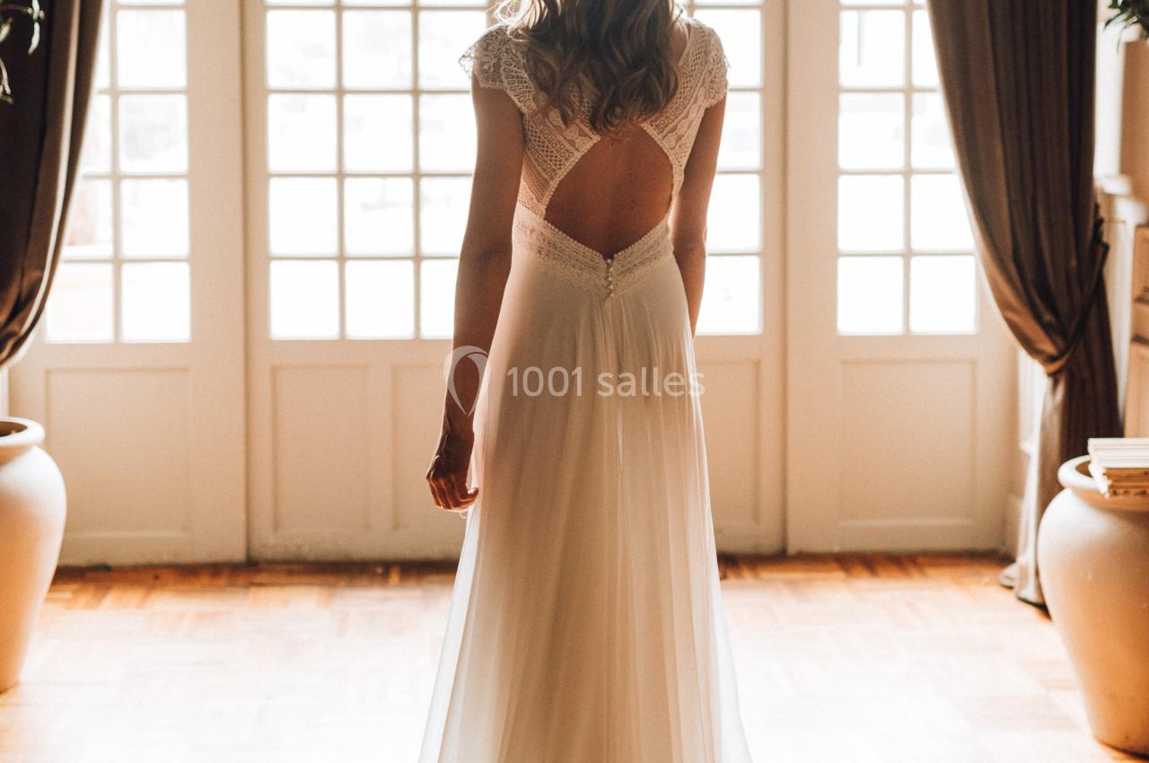 Femme en robe de mariée blanche, vue de dos, devant une grande baie vitrée avec rideaux sombres.