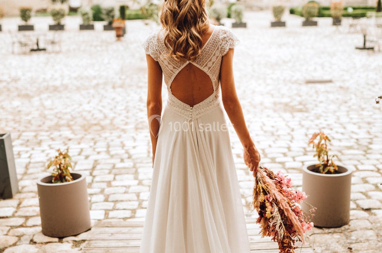 Une femme en robe de mariée blanche, vue de dos, tenant un bouquet de fleurs séchées devant un bâtiment en pierre.