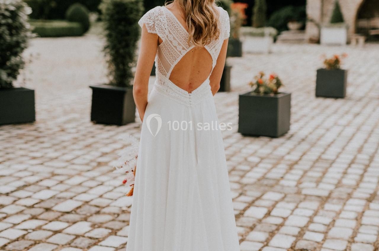 Femme en robe de mariée blanche vue de dos, debout sur une allée pavée entourée de verdure et de pots de fleurs.