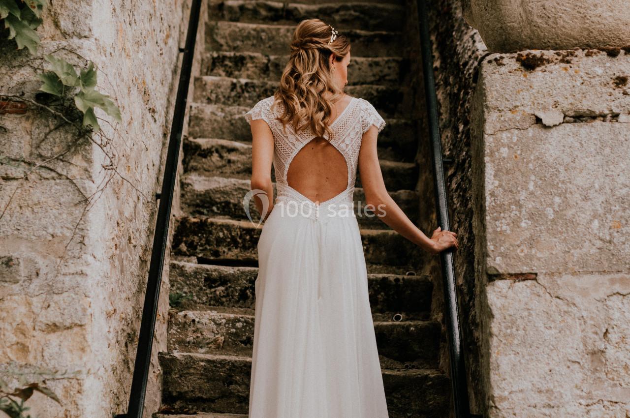 Femme en robe de mariée blanche montant un escalier en pierre, dos tourné, entourée de murs anciens et de végétation.