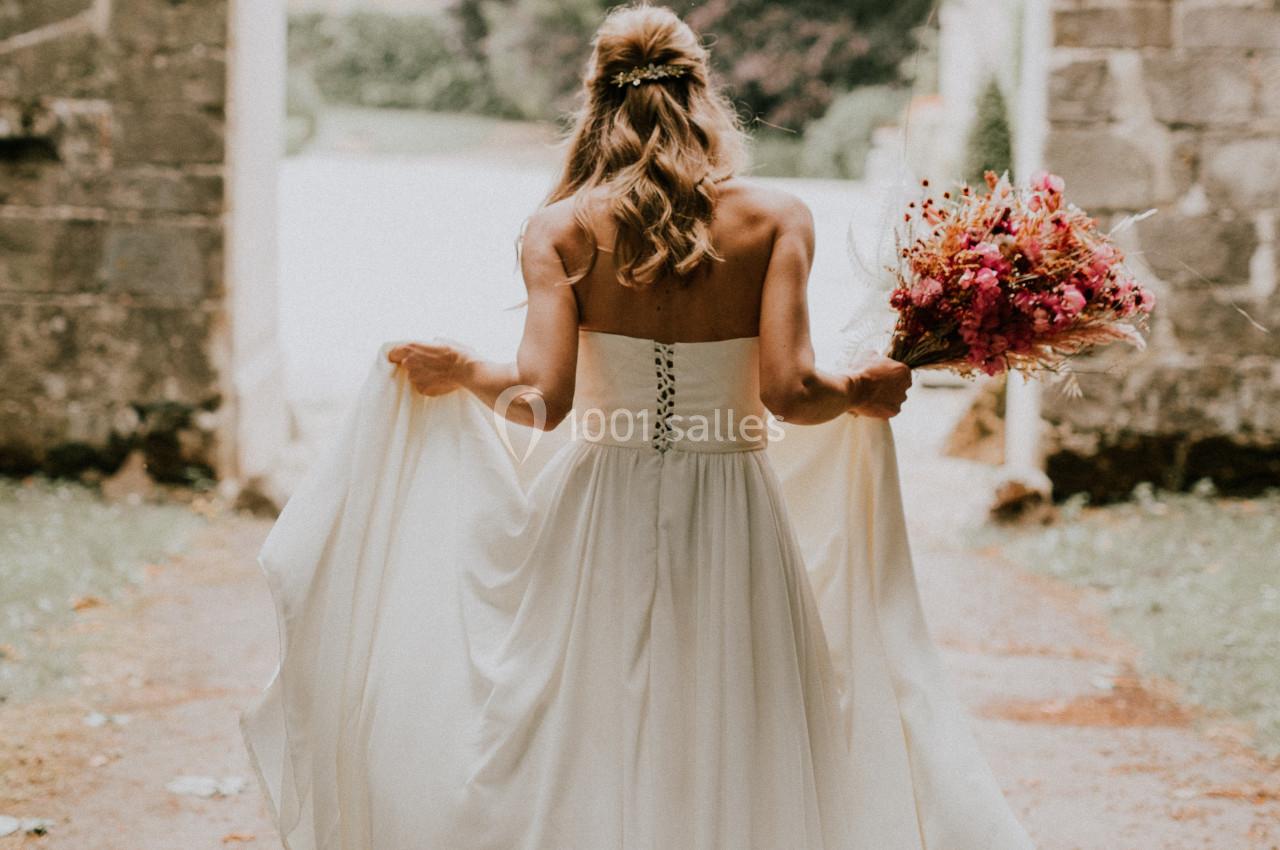 Une femme en robe de mariée blanche tient un bouquet coloré en marchant dans une allée bordée de pierres.