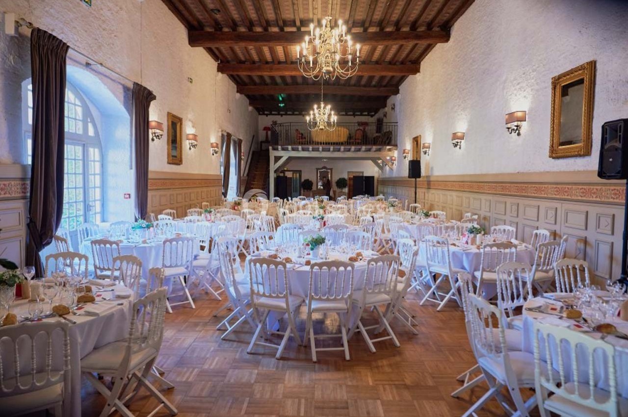 Salle de réception élégante avec tables rondes dressées, chaises blanches et lustres suspendus sous un plafond en bois.