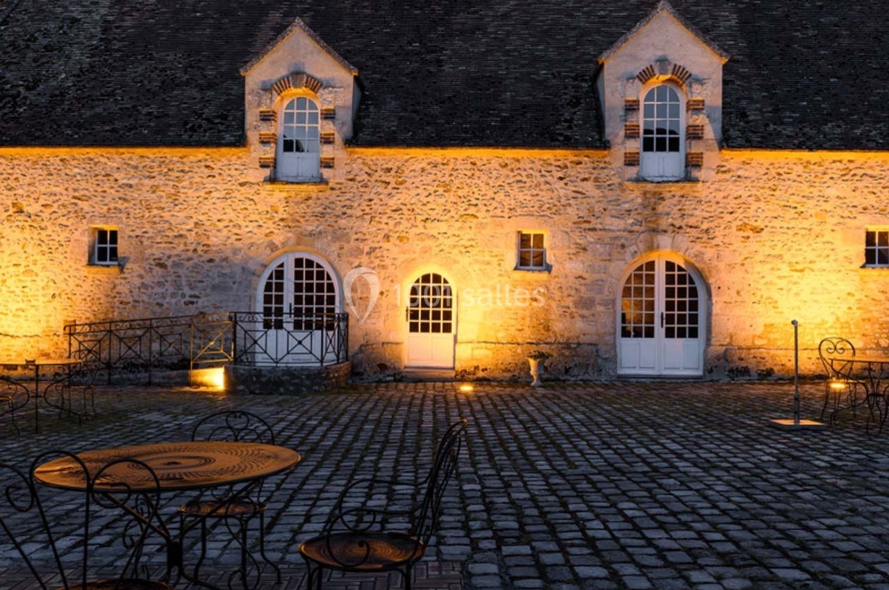 Façade en pierre d'un bâtiment ancien éclairé la nuit, avec tables et chaises en fer forgé sur une cour pavée.