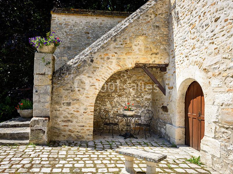 Cour en pierre avec table et chaises en fer forgé sous un escalier, entourée de murs en pierre et de fleurs.