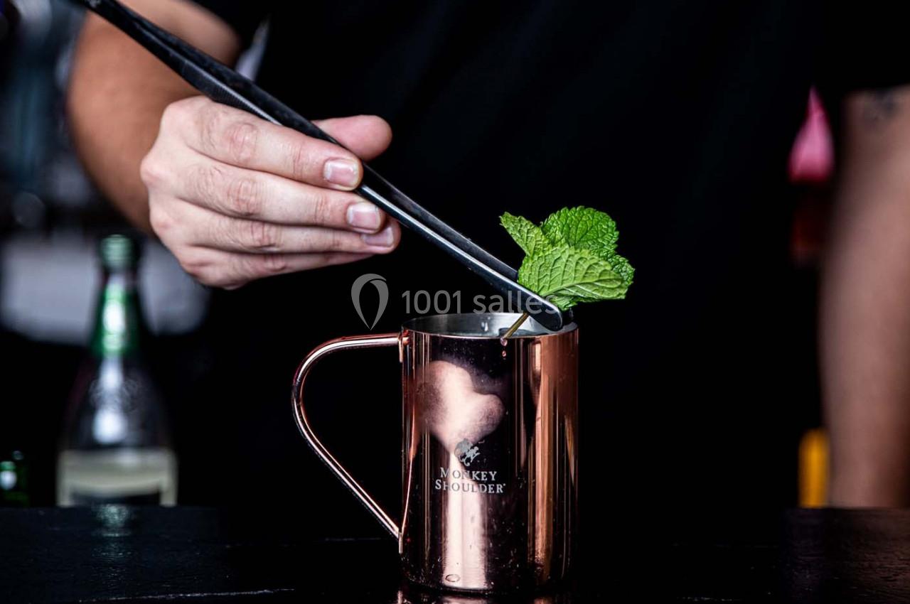 Une main utilise des pinces pour placer une feuille de menthe dans un mug en métal cuivré sur un comptoir.