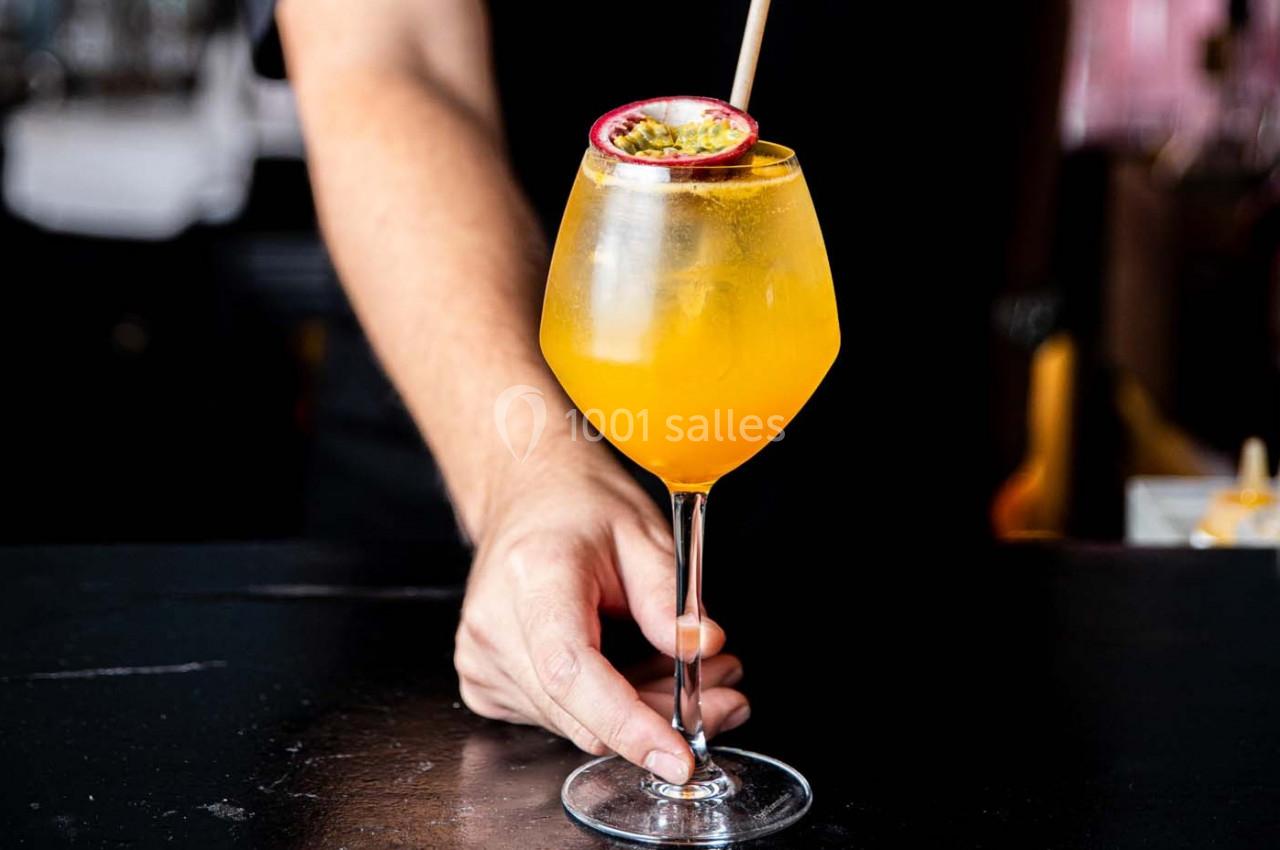 Verre à cocktail orange décoré d'un demi-fruit de la passion, tenu par une main sur un comptoir sombre.