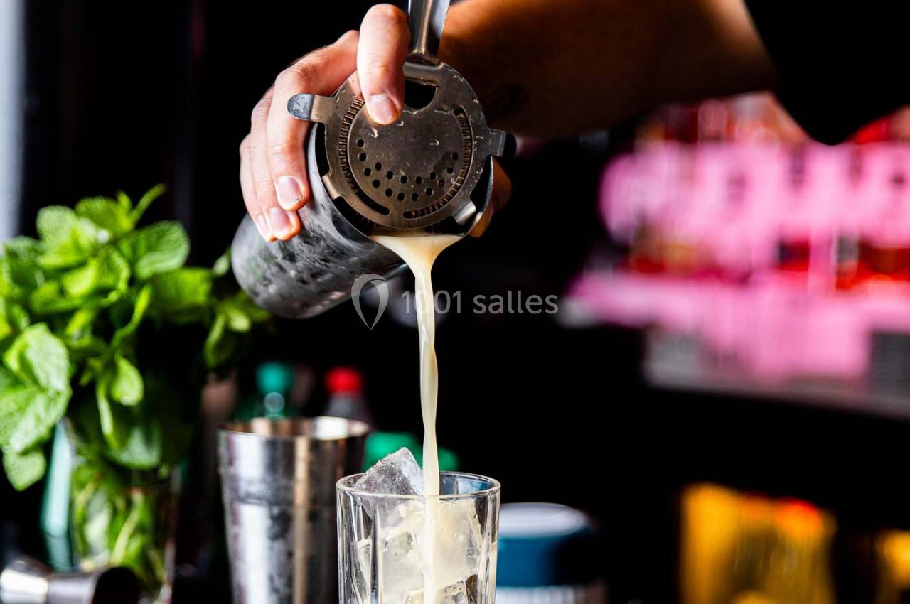 Un barman verse un cocktail filtré dans un verre rempli de glaçons, avec des herbes fraîches en arrière-plan.