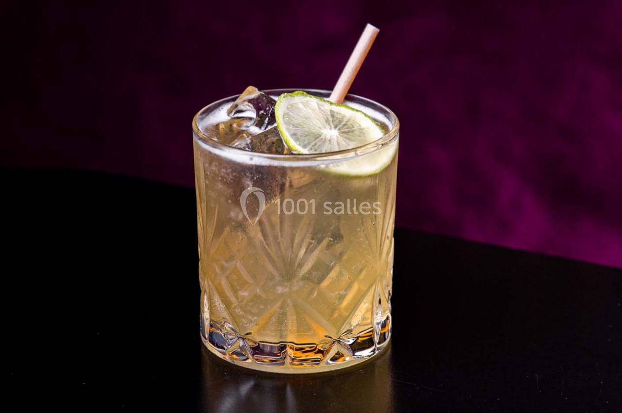 Verre de cocktail avec glaçons, tranche de citron vert et paille, posé sur une surface noire avec un fond violet.