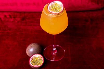 Cocktail jaune orangé garni d’un demi-fruit de la passion, accompagné d’un verre de champagne sur un comptoir en bois.
