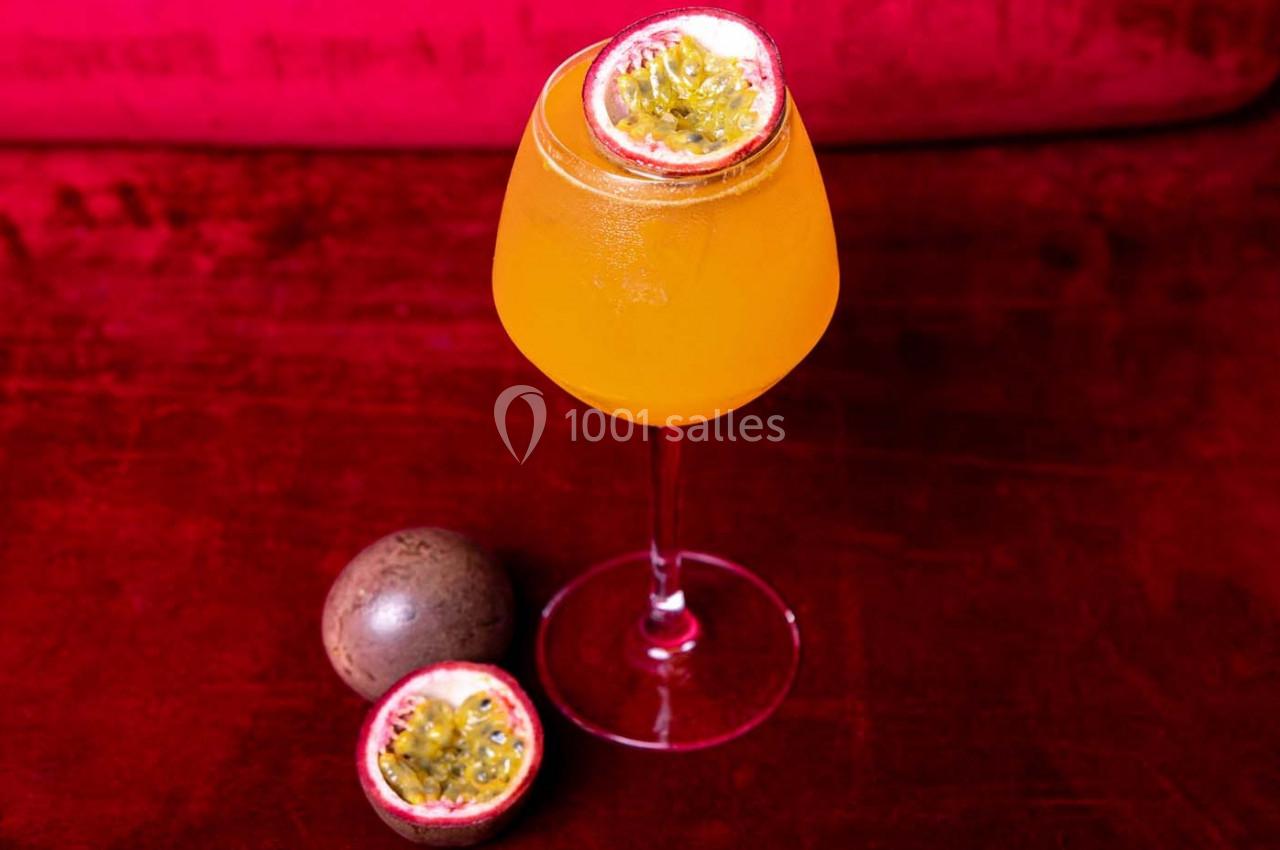Verre de cocktail orange garni d'une demi-passion, posé sur une table rouge avec deux fruits de la passion entiers.