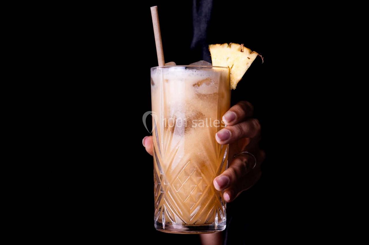 Main tenant un verre de cocktail décoré d'un morceau d'ananas, sur fond noir.