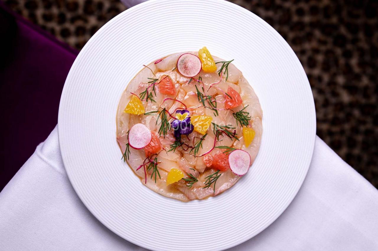 Assiette de carpaccio de poisson garnie d'agrumes, radis, herbes fraîches et une fleur comestible, vue de dessus.