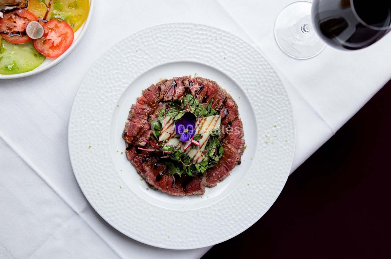 Assiette de carpaccio de bœuf garnie de légumes, herbes fraîches et fleur comestible, servie sur une nappe blanche.