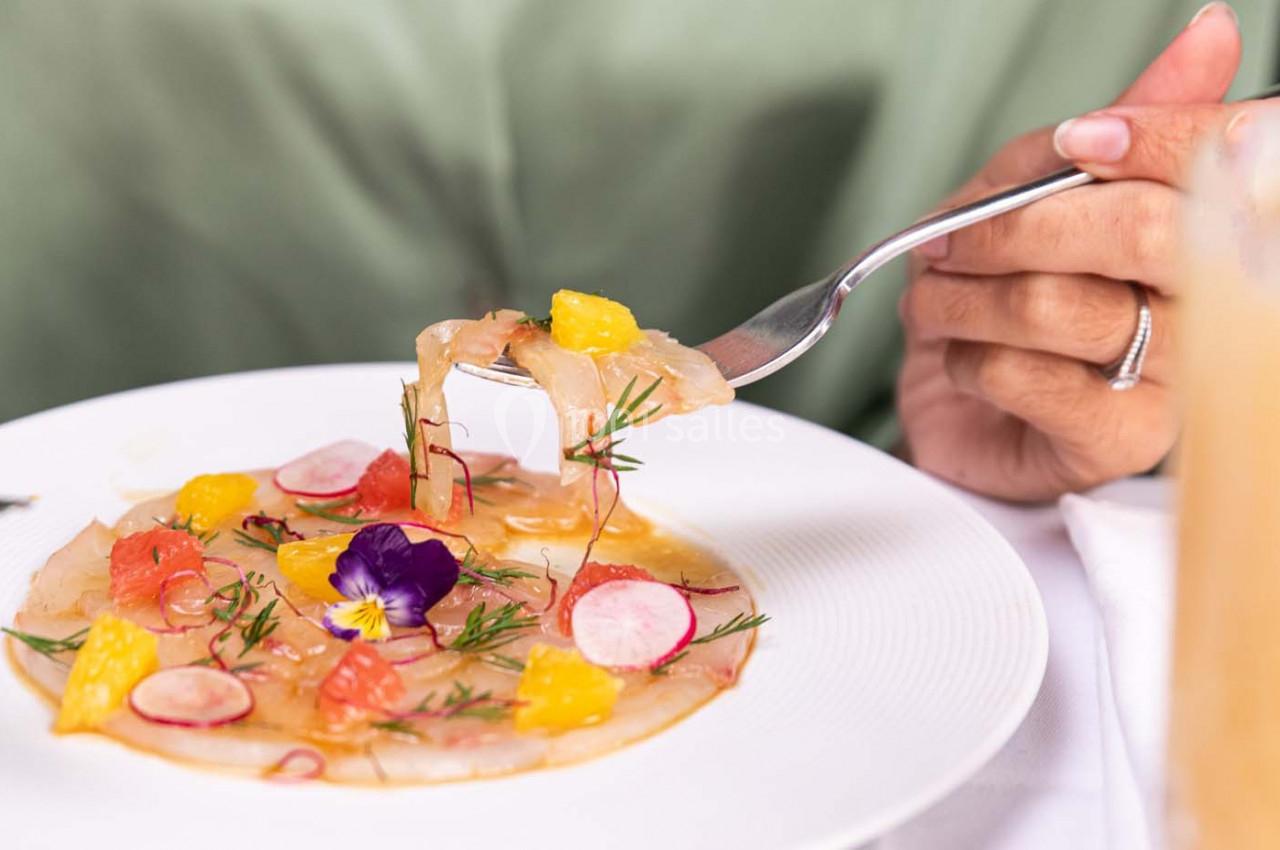 Assiette de carpaccio de poisson garnie de radis, agrumes, herbes fraîches et fleurs comestibles, tenue par une fourchette.