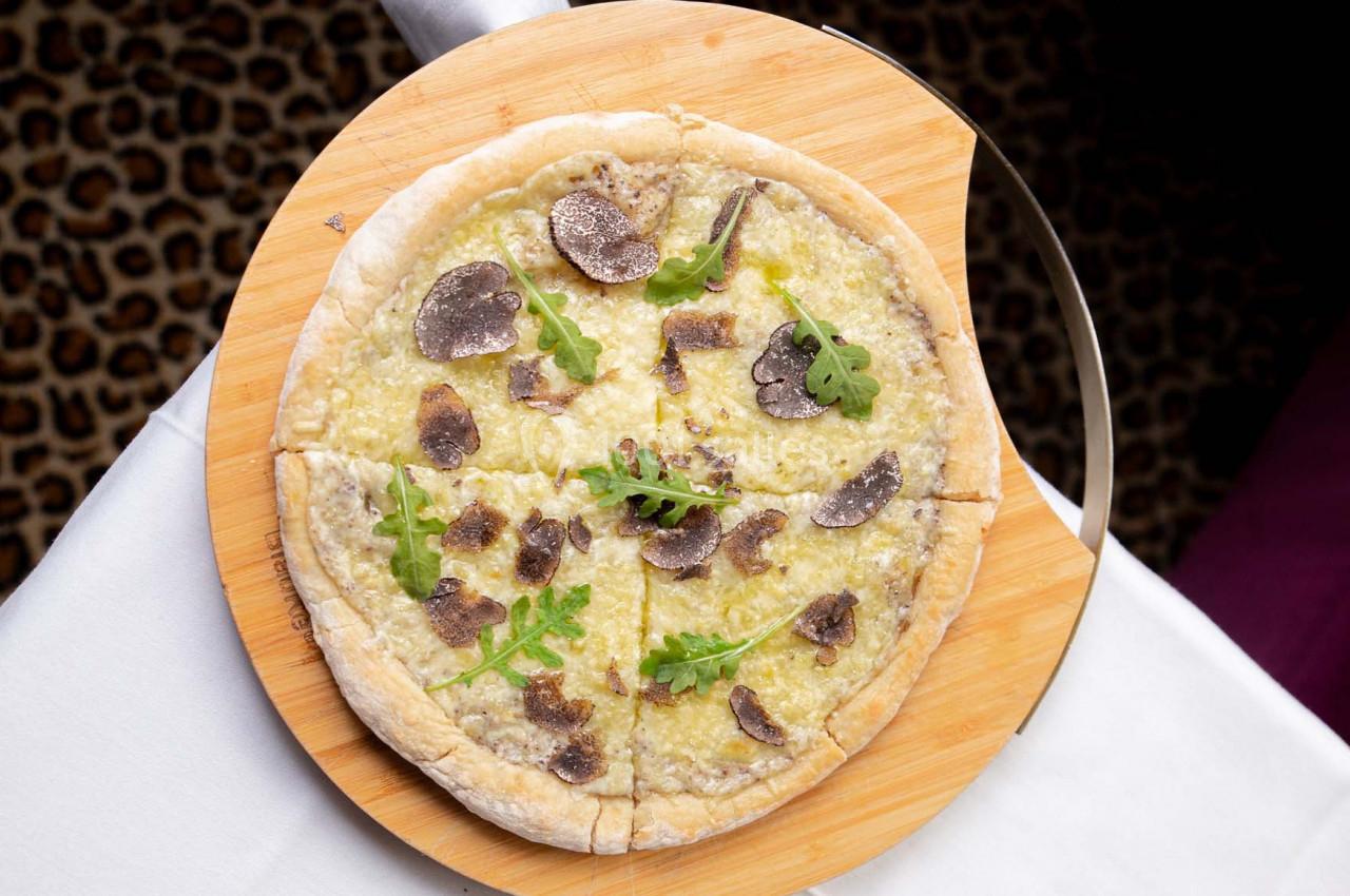 Pizza garnie de truffes, roquette et fromage, servie sur une planche en bois sur une table blanche.