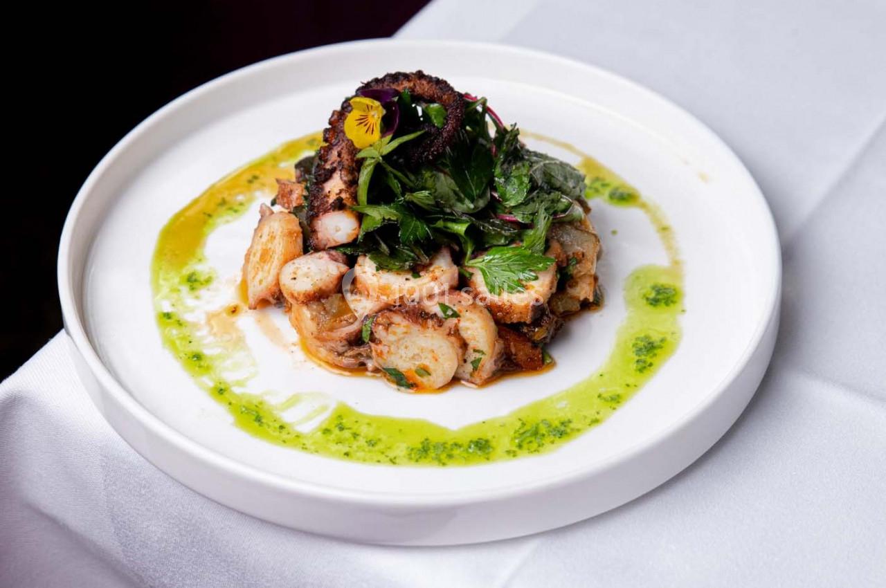 Assiette de poulpe grillé accompagné de pommes de terre, herbes fraîches et sauce verte, présentée sur une nappe blanche.