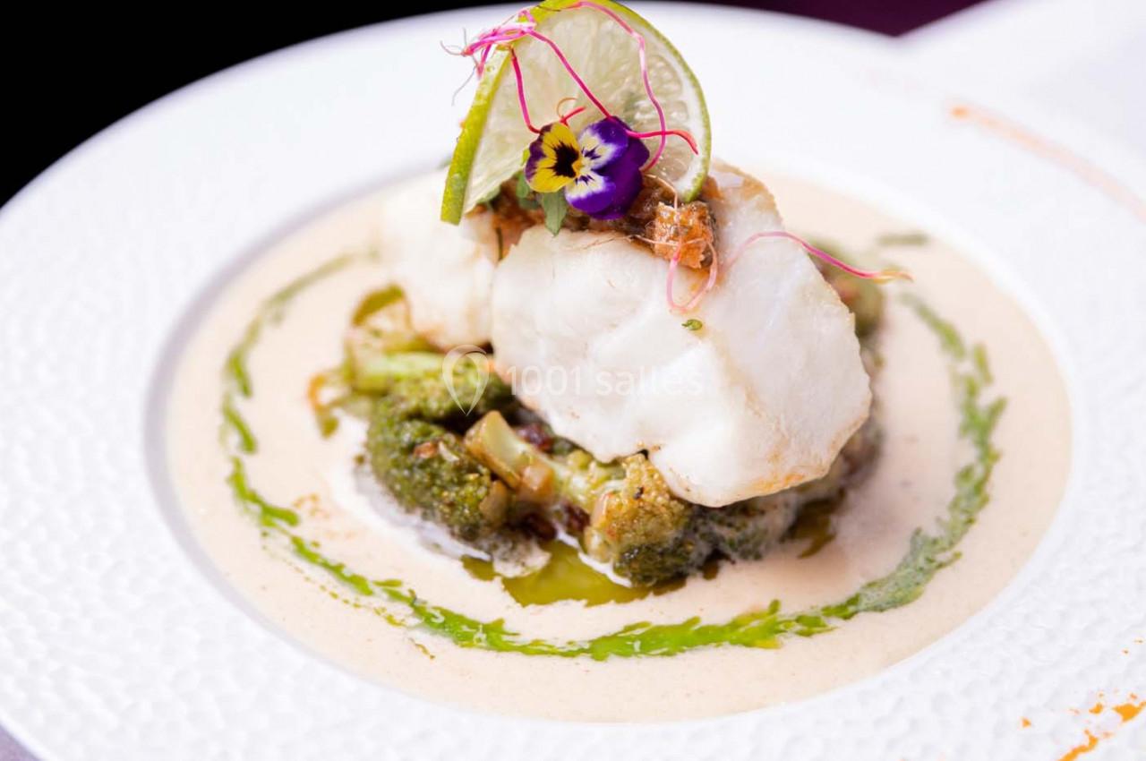 Plat de poisson blanc sur lit de brocolis, nappé de sauce crémeuse, décoré de fleurs comestibles et d'une tranche de citron…