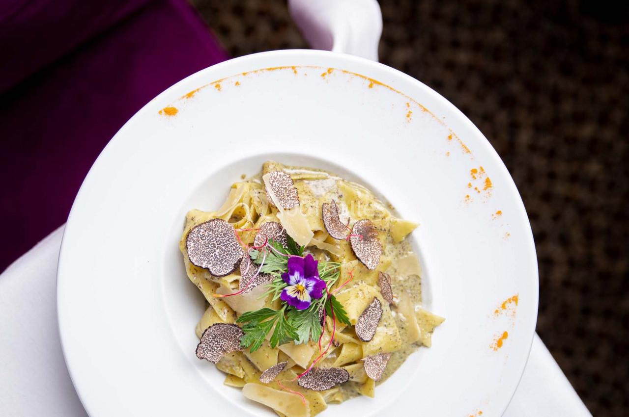 Assiette de pâtes garnie de truffes, fleurs comestibles et herbes, présentée sur une nappe blanche.