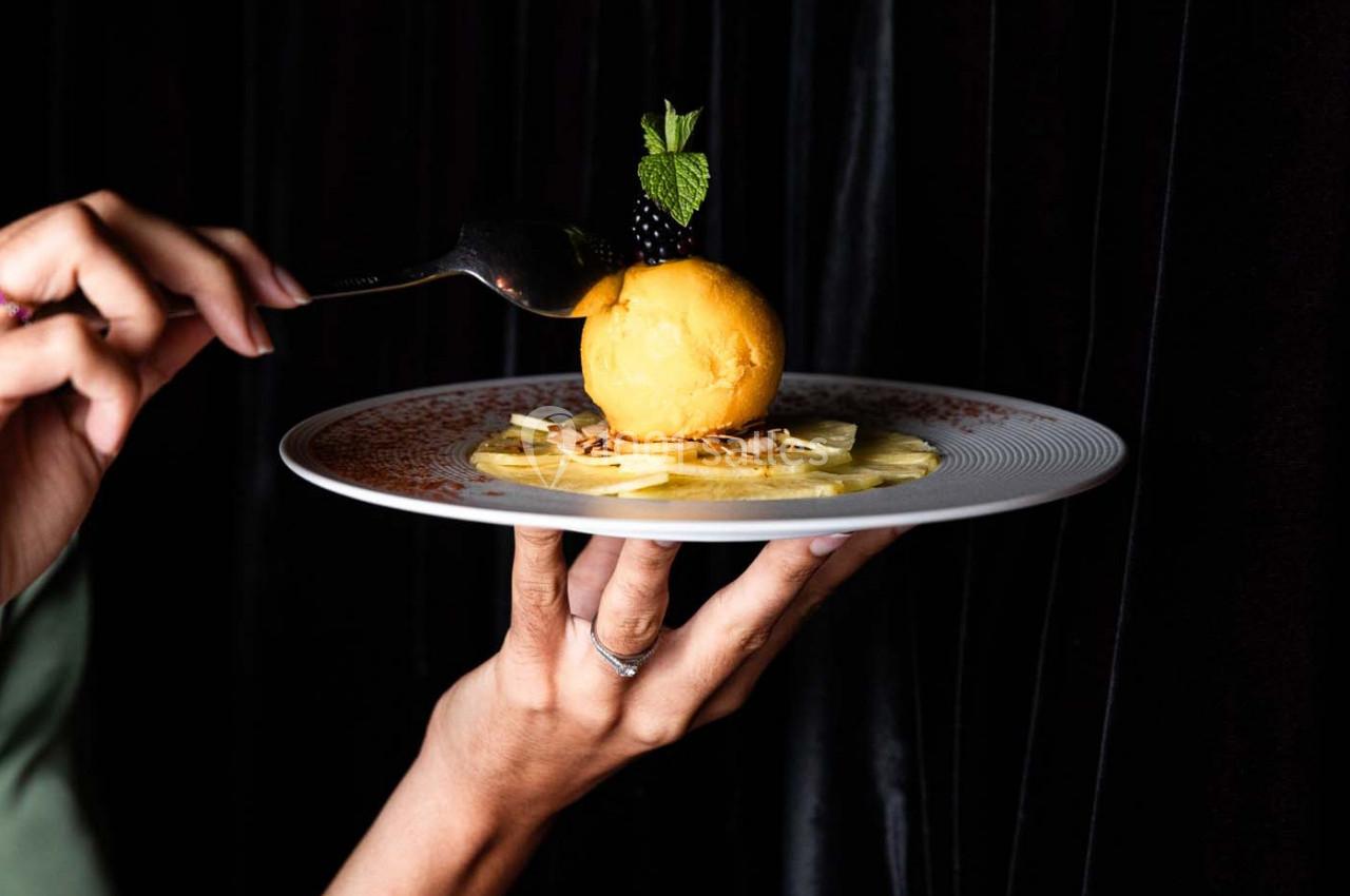 Une main tient une assiette avec une boule de sorbet sur des tranches d'ananas, décorée de mûres et de menthe.