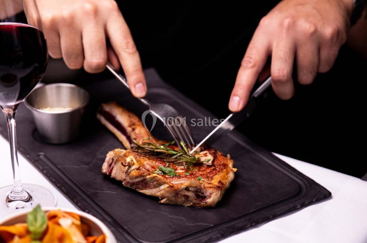 Une personne découpe une côte de viande grillée garnie de romarin sur une assiette noire, avec un verre de vin rouge à côté.