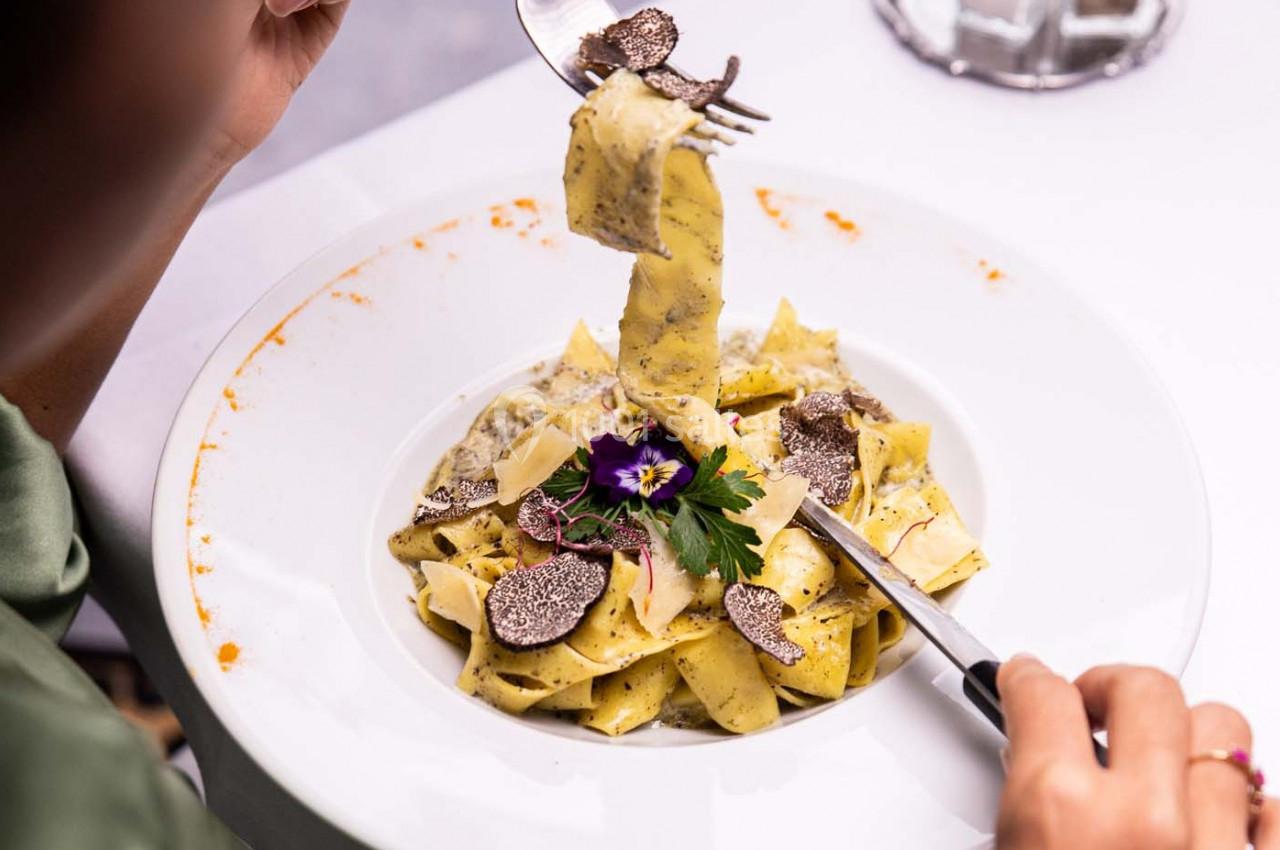 Assiette de pâtes garnies de truffes, copeaux de fromage et décorée d'une fleur comestible, sur une table blanche.