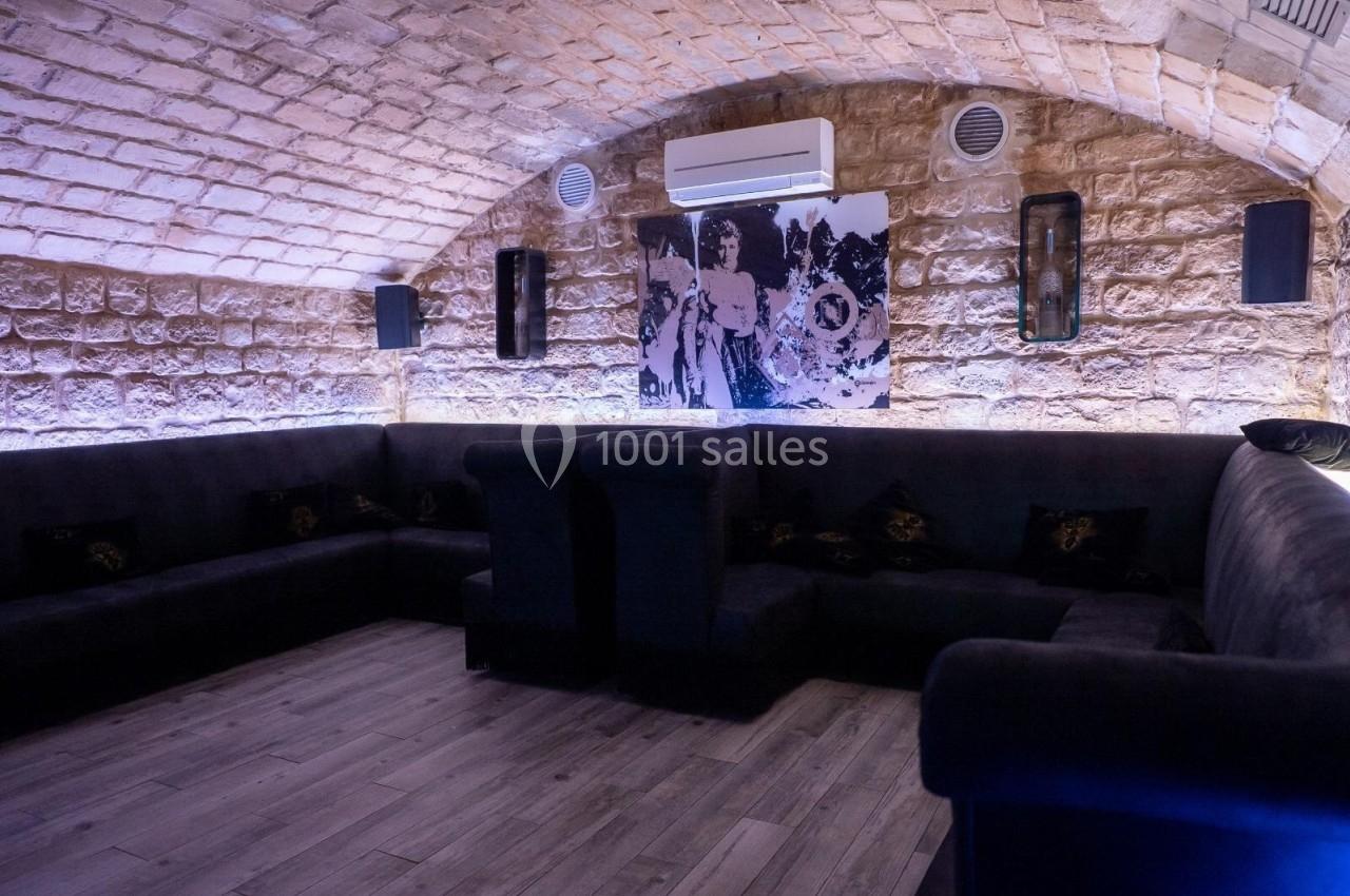 Salle voûtée en pierre avec canapés sombres, éclairage tamisé et tableau décoratif au mur.