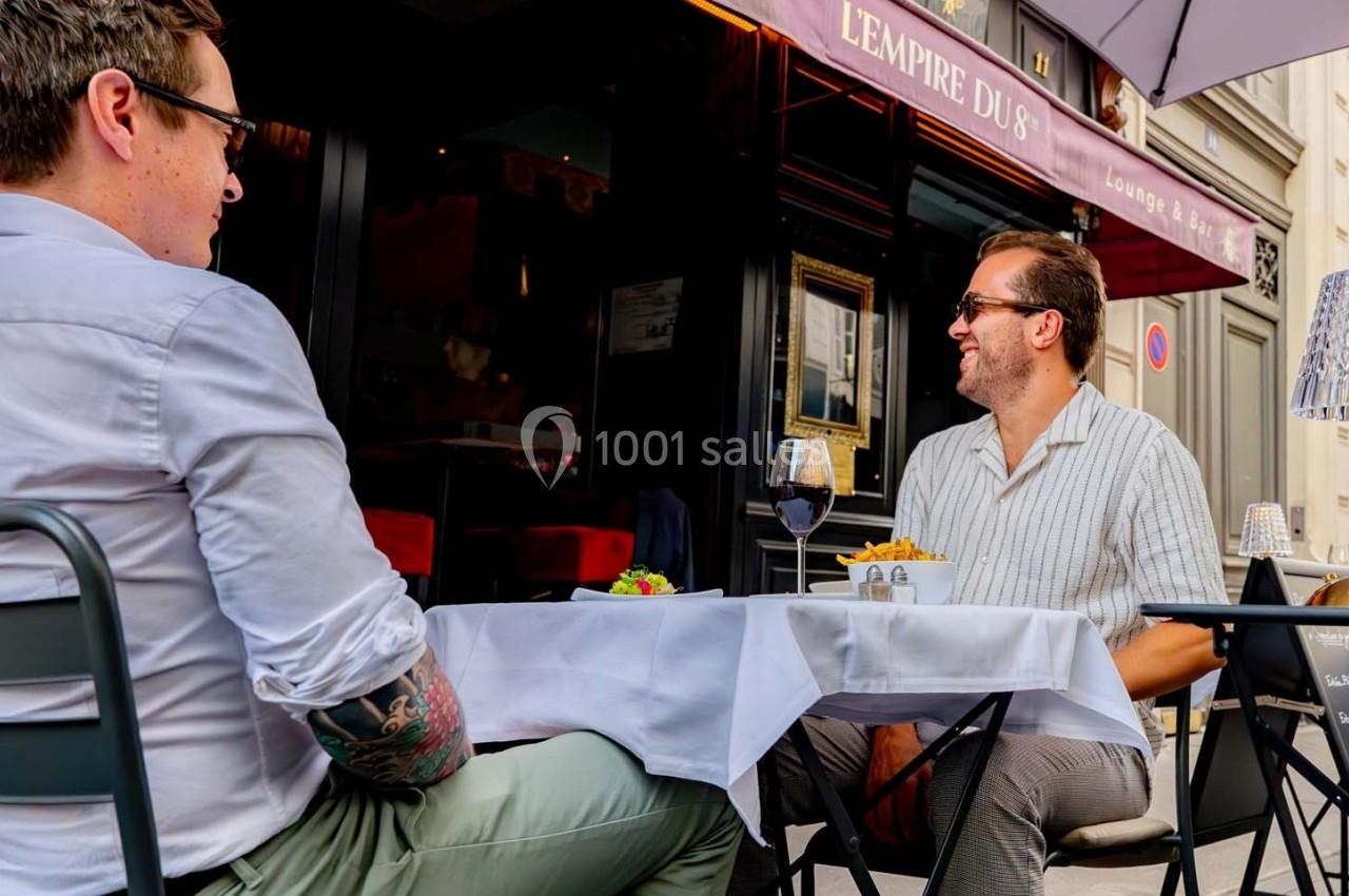 Deux hommes discutent à une table en terrasse d'un restaurant, avec un verre de vin et des frites sur la table.