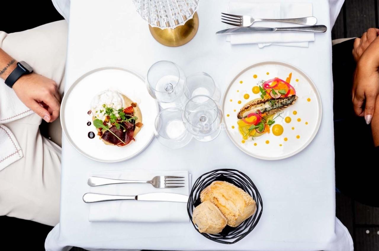 Vue aérienne d'une table dressée avec deux assiettes gastronomiques, des couverts, des verres et un panier de pain.
